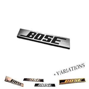 May include: A rectangular metal emblem featuring the word BOSE in bold black lettering. The primary piece has a brushed silver finish. Additional variations shown below include metallic finishes in rose gold, dark grey, and gold with black text.