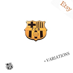 Op de afbeelding: Een schildvormig embleem met het FCB-logo in zwart en goud, voorzien van een kruis, verticale strepen en een voetbalafbeelding. Het ontwerp bevat de letters FCB in het midden. Een kleinere monochrome versie is onderaan zichtbaar.