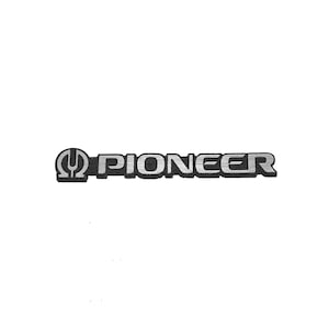 Può includere: Un'emblema "PIONEER" color argento con un logo stilizzato a sinistra. L'emblema ha un aspetto in metallo spazzolato ed è posto su uno sfondo bianco. Il testo è in un font sans-serif.