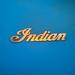 Può includere: Un'insegna in metallo color oro con la parola Indian in un carattere corsivo stilizzato. Le lettere presentano una texture spazzolata e un contorno scuro, creando un netto contrasto con lo sfondo blu solido e vibrante.