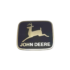 Könnte beinhalten: Ein rechteckiges Emblem mit einem goldfarbenen Hirsch, der nach rechts springt, vor einem dunkelblauen Hintergrund. Die Worte "JOHN DEERE" stehen in weißen Blockbuchstaben unter dem Hirsch, mit einem goldenen Rand.