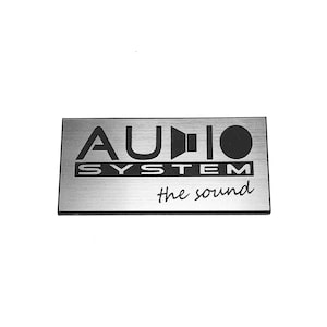 Könnte beinhalten: Ein rechteckiges, silberfarbenes Metallschild mit gebürsteter Oberfläche. Der Text lautet AUDIO SYSTEM in schwarzen Großbuchstaben, wobei ein Lautsprechersymbol den Buchstaben D ersetzt. Darunter steht der Schriftzug the sound in schwarzer Schreibschrift.