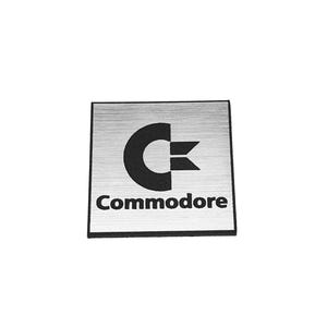 May include: A square, brushed silver-colored metal plate featuring a black Commodore logo and the word Commodore printed in black text. The plate has a thin black border around its perimeter, providing a clean, high-contrast visual design.