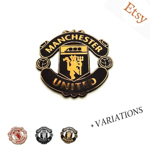 Puede incluir: Un pin metálico circular con el escudo del club de fútbol Manchester United en negro y dorado. El diseño incluye el nombre del equipo, un barco y una figura de diablo con un tridente. Tres variaciones más pequeñas se muestran debajo.