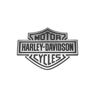 May include: A silver and black metal emblem featuring the Harley-Davidson Motor Cycles shield logo. The design includes a central horizontal bar with the text Harley-Davidson in black, flanked by the words Motor and Cycles in a shield shape.