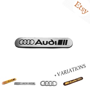 May include: A rectangular silver-toned metal emblem featuring the black Audi logo and four rings, with additional variations shown below including gold-toned, black, and silver-toned badges with text like Limited Edition and the Audi logo.