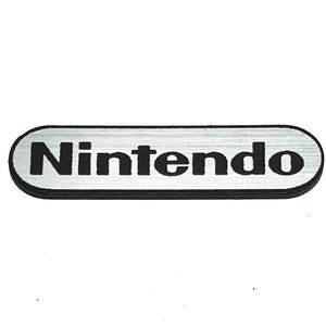 May include: A rectangular, horizontally oriented badge with rounded ends, featuring a brushed silver metallic finish and a black border. The word Nintendo is printed in bold, black, sans-serif capital letters across the centre of the badge.