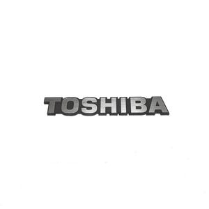 Può includere: Un emblema metallico argentato con il logo Toshiba, caratterizzato da una texture spazzolata sulla superficie delle lettere. La scritta è in grassetto e in maiuscolo, su un contorno scuro e contrastante che definisce il nome del marchio.
