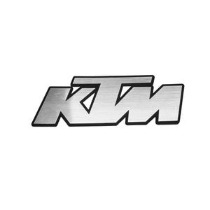 Könnte beinhalten: Ein silberfarbenes Emblem aus Metall mit den Buchstaben KTM in einer fetten, stilisierten Schriftart. Die Buchstaben haben eine gebürstete Metalloberfläche und sind mit einem dünnen schwarzen Rand versehen, der einen Kontrast bildet.