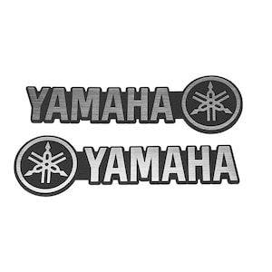 May include: Two metallic Yamaha emblems featuring a brushed silver finish on the lettering and the circular logo. Each emblem displays the word YAMAHA in bold capital letters next to a circular icon containing three crossed tuning forks.