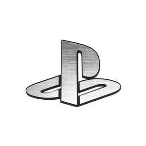 以下が含まれることがあります： ブラッシュ仕上げの質感を備えたメタリックシルバーのPlayStationロゴエンブレム。この象徴的なシンボルは、垂直の直立バーと湾曲した水平ベースで構成されており、どちらも白い背景に対して細い黒い境界線で縁取られています。