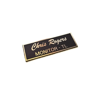 Puede incluir: Una placa de identificación rectangular con una superficie negra texturizada y un borde dorado. El nombre Chris Rogers está grabado en fuente cursiva y el texto MONITOR - TL está grabado en una fuente sans-serif, revelando una base dorada.