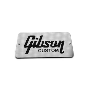 May include: A rectangular brushed metal plate featuring the black Gibson logo with a star and the word CUSTOM underneath. The plate has rounded corners and two small black screws on the left and right sides for mounting purposes.