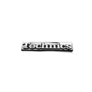 Könnte beinhalten: Ein rechteckiges Metall-Emblem mit dem Wort Technics in erhabenen, silberfarbenen Buchstaben auf schwarzem Hintergrund. Das Logo hat eine polierte, reflektierende Oberfläche mit klarer, fetter Typografie und einem kleinen runden Detail über dem i.