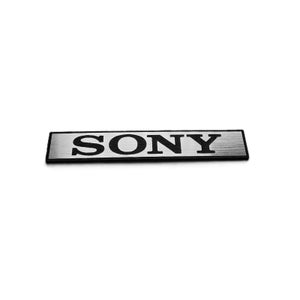 May include: A rectangular, silver metal emblem featuring the word "SONY" in black, sans-serif font. The emblem has a brushed metal texture and a black border. The logo is centered on a white background.