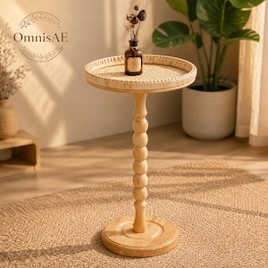 May include: A round wooden side table with a woven rattan tray top and a central pedestal base featuring a stacked ball design. A small brown glass bottle containing dried botanical stems sits on the table surface. The brand name OmnisAE is visible.