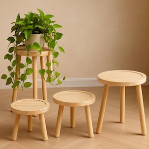 May include: A set of four light-colored wooden stools with round tops and three cylindrical legs. The stools vary in height, with the tallest one supporting a light-colored pot containing a trailing green plant with heart-shaped leaves.