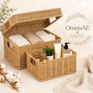 May include: Two rectangular woven storage baskets made of natural tan fibers. The larger basket has an open lid and contains three folded white and beige towels. The smaller basket holds two glass perfume bottles and a small potted green plant.