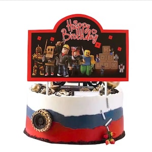 May include: A round cake with white, blue, and red frosting layers, decorated with a black tire, silver sprinkles, and a large rectangular cake topper featuring the text Happy Birthday and various block-style character figures in a gaming theme.