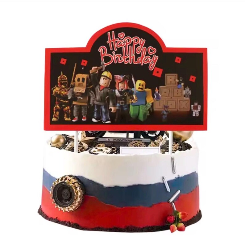 May include: A round cake with white, blue, and red frosting layers, decorated with a black tire, silver sprinkles, and a large rectangular cake topper featuring the text Happy Birthday and various block-style character figures in a gaming theme.