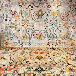 May include: A large area rug featuring a cream-colored background with an intricate, symmetrical pattern of floral and botanical motifs in shades of red, yellow, blue, green, and orange. The design includes a decorative border and fringe.