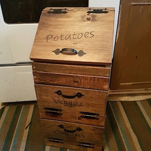 May include: A wooden storage bin with three drawers labelled "Potatoes", "Veggies", and "Onions". Each drawer has a black metal handle and decorative hinges.