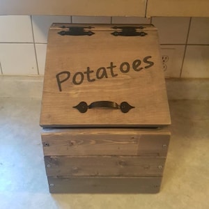 May include: A wooden storage bin with a hinged lid and metal handles. The bin is painted a light brown color and has the word "Potatoes" written on the front.