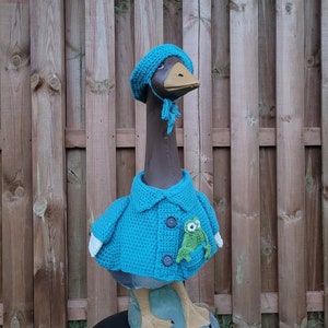 Puede incluir: Una estatua de ganso lleva un gorro azul tejido y una chaqueta a juego. El ganso lleva una pequeña rana verde en su bolsillo.