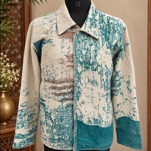 Puede incluir: Una chaqueta de manga larga con una superficie texturizada y acolchada, con una base de color crema y un patrón abstracto en azul cerceta y marrón apagado. La prenda tiene un cuello estándar, cierre de botones y costuras visibles.