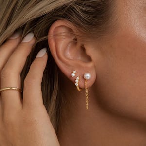 May include: A close-up of a person's ear adorned with three gold earrings. One earring features a pearl, another has a chain, and the third has small stones. A gold ring is on the finger.