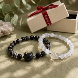 May include: Two beaded bracelets featuring round stones and a central cat-shaped charm. One bracelet consists of dark, polished black beads, while the other features translucent white beads. Both bracelets include small silver-toned metal spacers.