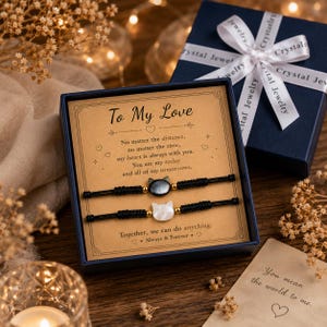 Könnte beinhalten: Ein Geschenkset mit zwei schwarzen Kordelarmbändern mit katzenförmigen Anhängern, eines schwarz und eines weiß, präsentiert in einer marineblauen Schachtel mit einer Karte mit der Aufschrift "To My Love". Die Schachtel ist mit einem weißen Band gebunden.