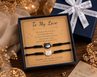 Puede incluir: Un juego de regalo con dos pulseras de cordón negro con dijes en forma de gato, uno negro y otro blanco, presentado en una caja azul marino con una tarjeta que dice "To My Love". La caja está atada con una cinta blanca.