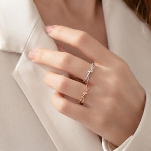Könnte beinhalten: Zwei dünne Ringe an den Fingern, einer in Silberoptik und einer in Roségoldoptik. Jeder Ring verfügt über ein zartes, gekreuztes Knotendesign in der Mitte, das mit kleinen, klaren, funkelnden Steinen entlang der Ringschiene besetzt ist.