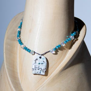Può includere: Una collana fatta a mano con un ciondolo raffigurante un edificio. Il ciondolo è bianco con dettagli verdi e grigi. La collana ha una catena di perline blu con accenti di cristallo trasparente. La collana è esposta su un busto di legno.
