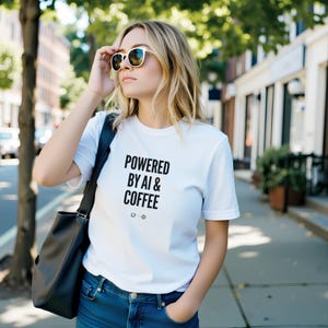 May include: A white short-sleeved t-shirt featuring the black text "POWERED BY AI & COFFEE" with small icons of a coffee cup and a snowflake. The shirt is paired with blue denim jeans and a black shoulder bag, worn with dark-rimmed sunglasses.