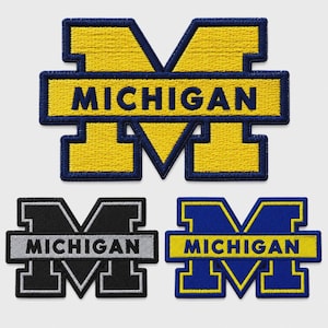 May include: Three embroidered patches featuring the block letter M with the word MICHIGAN centered across the middle bar. The top patch is yellow with blue borders, the bottom left is black with grey borders, and the bottom right is blue with yellow borders.