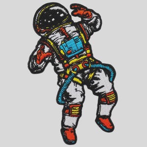May include: An embroidered patch featuring an astronaut in a white suit with black, yellow, red, and blue accents. The astronaut is depicted in a floating pose with a black helmet visor, a blue chest pack, and colorful details on the limbs and gear.