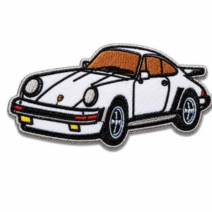 May include: An embroidered patch featuring a white sports car with a distinctive sloping roofline, orange-tinted windows, black trim, and a rear spoiler. The design includes circular headlights, yellow fog lights, and a small emblem on the hood.