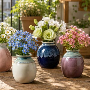 Puede incluir: Cuatro jarrones de cerámica pequeños y redondos en tonos rosa, blanco, azul oscuro y morado, cada uno con un acabado esmaltado liso. Los jarrones están llenos de varias flores frescas, incluyendo margaritas blancas, nomeolvides azules y flores rosas.