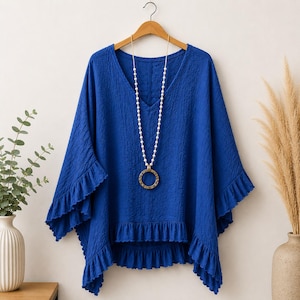 May include: A vibrant royal blue V-neck tunic top featuring a textured fabric, wide sleeves with ruffled edges, and a matching ruffled hemline. The garment is displayed on a wooden hanger and is accessorised with a long necklace featuring white beads.