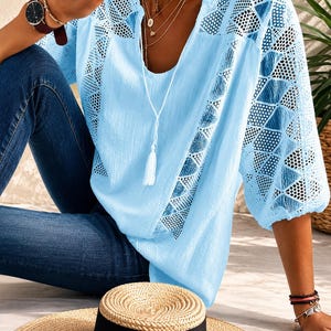Può includere: Una camicetta azzurra con scollo a V e laccetto con nappa bianca, maniche a tre quarti e pannelli in pizzo geometrico lungo le spalle e i fianchi. Il tessuto ha una finitura testurizzata ed è abbinato a jeans blu scuro.