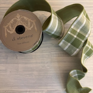 Op de afbeelding: Een spoel groen lint met de tekst "d. stevens fine ribbons" op het etiket. Het lint heeft een effen groene kleur en een groen-wit ruitpatroon. Het lint ligt op een licht houten oppervlak.