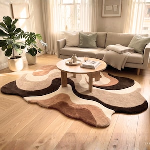 May include: An irregular, organic-shaped area rug featuring a layered, wavy pattern in shades of cream, beige, tan, and dark brown. The rug has a thick, tufted texture and is positioned on a light-colored wooden floor beneath a round table.