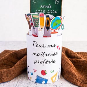Può includere: Un portapenne cilindrico bianco con la scritta "Pour ma maîtresse préférée" e illustrazioni a tema scolastico. Presenta una piccola lavagna con "Année 2025-2026" e ritagli di materiale scolastico.