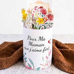 Op de afbeelding: Een witte cilindrische geschenkdoos met een bloemenontwerp en de tekst "Pour Ma Maman que j'aime fort". De doos is gevuld met papieren bloemen en een envelop. Een bruine doek is op de achtergrond gedrapeerd.