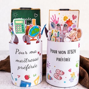 Puede incluir: Dos recipientes cilíndricos blancos decorados con ilustraciones coloridas de temática escolar. El recipiente izquierdo lleva el texto "Pour ma maîtresse préférée" y el derecho "POUR MON ATSEM PRÉFÉRÉE", ambos llenos de recortes de papel.