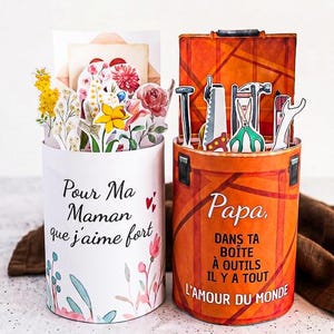 May include: Two decorative paper containers. One white with floral accents and the text "Pour Ma Maman que j'aime fort". The other is orange, resembling a toolbox, with tools and the text "Papa, Dans ta boîte à outils il y a tout l'amour du monde".