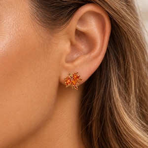 May include: A stud earring shaped like a maple leaf, featuring multiple orange and amber-colored gemstones set in a gold-toned metal frame. The leaf design has five distinct points and is positioned on the earlobe, showcasing a vibrant autumn color.