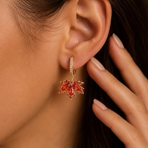May include: A gold hoop earring featuring a dangling maple leaf charm. The hoop is encrusted with small clear stones, while the maple leaf charm is composed of multiple marquise-cut orange gemstones arranged in a vibrant, autumnal leaf pattern.
